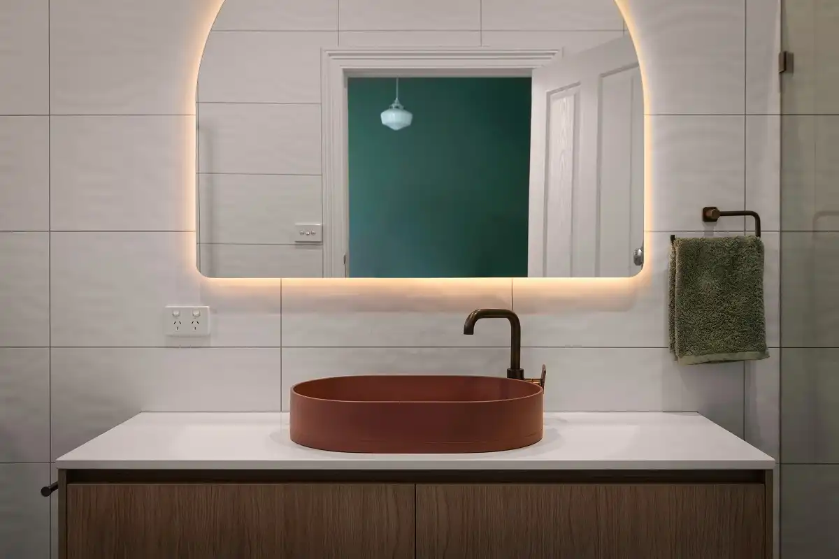 Bathroom vanity with a rust vessel sink, black faucet, arched backlit mirror, and white tiles with dark contrasting grout.